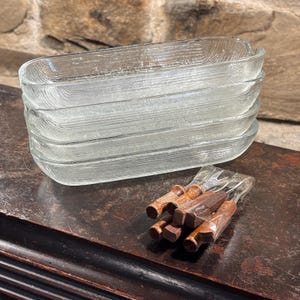 May include: Four clear, rectangular glass dishes with a textured surface are stacked. A bundle of wooden skewers with brown and reddish-brown tones is in the foreground. The items are on a dark wooden surface.