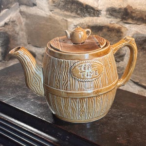 Pode incluir: Uma chaleira de cerâmica marrom com um design de barril e as palavras "TEA TIME" na frente. A chaleira tem uma pequena tampa em forma de chaleira e uma alça curva. A chaleira é feita de cerâmica e tem uma aparência rústica.