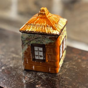 May include: A ceramic sugar bowl shaped like a cottage, with a thatched roof, brown brick walls, and a window with white panes. The bowl is decorated with green vines and flowers.