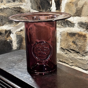 May include: A tall, cylindrical, amethyst-coloured glass vase with a wide, flat rim. The vase features a circular, embossed floral design near the base. The vase is displayed on a dark wooden surface against a stone wall.
