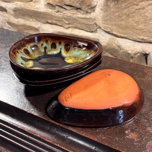 May include: A set of teardrop-shaped ceramic dishes with a dark brown exterior and a glossy interior glaze featuring blended tones of deep brown, teal, and yellow. One dish is inverted to show the unglazed terracotta-colored base with a maker's mark.