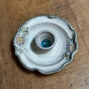 May include: A handcrafted ceramic ashtray with a central cup and floral detailing. The ashtray is off-white with a green edge, and a blue glass bead sits in the cup. The floral elements are in green, yellow, and blue hues.