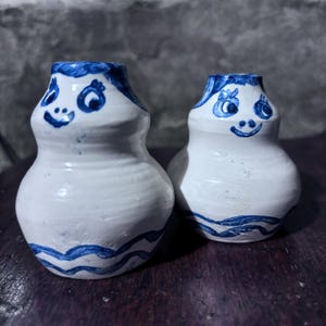 Pode incluir: Dois vasos de cerâmica branca com detalhes pintados em azul. Cada vaso tem um design de rosto sorridente com olhos azuis e uma linha ondulada azul na base. Os vasos são feitos à mão e têm um estilo de arte popular caprichoso.