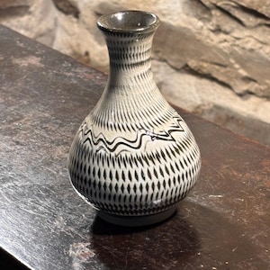 Puede incluir: Un jarrón decorativo de cerámica con cuello estrecho y cuerpo bulboso. El jarrón presenta un fondo color crema con patrones negros verticales y en forma de diamante, y un diseño de línea negra ondulada. El jarrón se exhibe sobre una superficie de madera oscura.