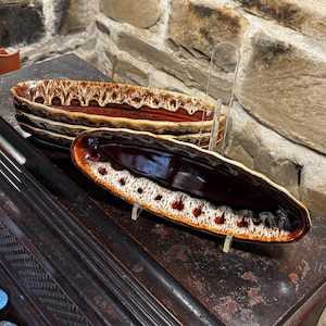 Puede incluir: Una pila de tres fuentes de cerámica marrón y blanca. Las fuentes, de forma ovalada, tienen un acabado brillante y un borde decorativo. El fondo muestra una pared de piedra y una superficie de madera oscura.