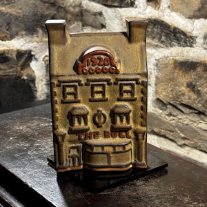 May include: A bronze-colored, rectangular decorative piece shaped like a building, with the text "THE BULL" at the bottom. The top of the building has the text "1920". The piece is mounted on a black base.