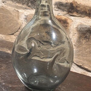 May include: A clear, teardrop-shaped glass bottle with a wide base and narrow neck. The bottle has a slightly smoky tint and a smooth surface. The background is a stone wall.