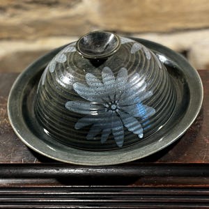 May include: A dark blue ceramic butter dish with a white floral design. The dish has a lid with a knob on top.