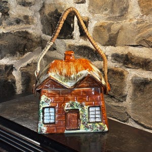 May include: A ceramic cottage-shaped container with a woven handle. The roof is brown and tan, and the walls are brick-patterned. Two windows and a door are visible, with green foliage accents. The handle is made of woven material.