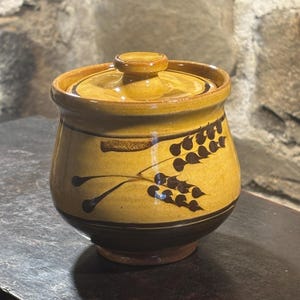 Puede incluir: Un tarro de cerámica con tapa, con un diseño de espiga de trigo. El tarro es principalmente amarillo mostaza con una base marrón oscuro y diseño de trigo. La tapa tiene un pequeño pomo. El tarro es probablemente hecho a mano y apto para la cocina.