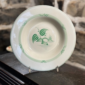 May include: A white ceramic soup bowl with a green rim and a green design of a bridge and foliage. The bowl is on a wooden surface.