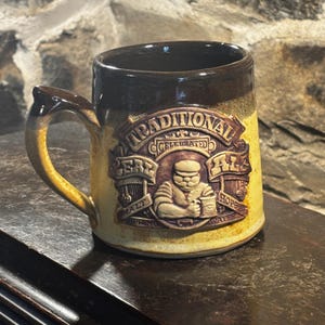 May include: A ceramic beer mug with a brown handle and rim, and a yellow body. The mug features a raised relief design with the words "Traditional Celebrated Ale" and an image of a man. The mug is sitting on a dark brown surface.