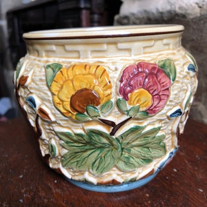 May include: A ceramic planter with a light brown base and a white glaze. The planter is decorated with two flowers, one yellow and one pink, and green leaves. The planter has a raised rim with a brown line.