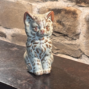 May include: A ceramic cat figurine featuring a speckled light green and brown glaze. The cat is in a seated position with pointed ears, defined facial features, and a textured coat pattern covering its entire body in a mottled brown finish.