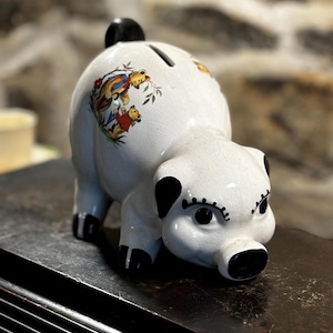 May include: White ceramic piggy bank with black eyes, eyelashes, and hooves. The piggy bank has a slot on the back and a painted design of three bears on the side.