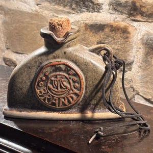 Puede incluir: Una jarra de cerámica con esmalte gris y marrón moteado, con un tapón de corcho y un emblema circular en relieve con el texto BRANDY y la ilustración de un barco. Un cordón de cuero negro está atado al asa y cuelga por el lateral del objeto.