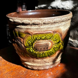 Puede incluir: Una maceta de cerámica marrón y verde con un diseño floral. La maceta tiene un borde elevado y una superficie texturizada.