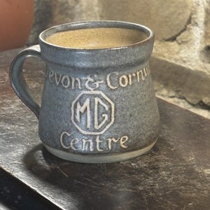 May include: A gray ceramic mug with a handle, featuring the text "Devon & Cornwall Centre" and an "MG" logo. The mug has a textured surface and a light brown interior, suggesting it's designed for hot beverages.