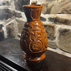 Könnte beinhalten: Eine braune Keramikvase mit aufwendigen floralen Reliefmustern. Die Vase hat einen breiten Sockel, einen bauchigen Körper und einen ausgestellten Hals. Der Hintergrund ist eine Steinmauer.