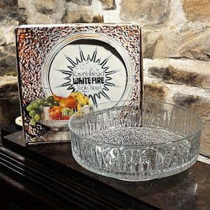 May include: A clear glass table bowl with a textured surface and a raised rim. The bowl is empty and is sitting on a wooden surface. The box for the bowl is in the background and says "Ravenhead Whitefire Table Bowl".