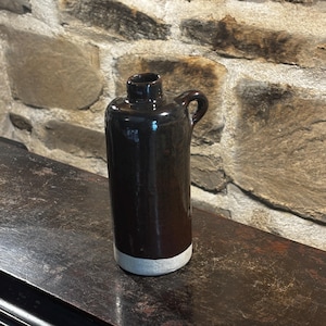 Op de afbeelding: Een donkerbruine keramische kan met een handvat en een witte basis. De kan heeft een cilindrisch lichaam en een kleine opening aan de bovenkant. De kan staat op een donker houten oppervlak.