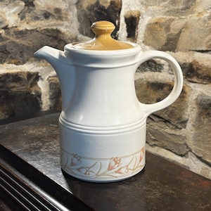 May include: White ceramic pitcher with a tan lid and handle. The pitcher has a floral design in peach and tan near the base. The pitcher is sitting on a dark wooden surface, with a stone wall in the background.