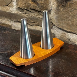 Puede incluir: Un juego de salero y pimentero. El juego presenta dos saleros y pimenteros cónicos de acero inoxidable en un soporte de madera. El soporte es rectangular con dos recortes circulares. La madera es de color marrón claro.