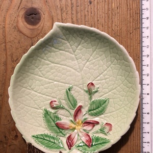 Op de afbeelding: Een groene, keramische schotel in de vorm van een blad met een reliëf van een bloem en bladeren. De bloem is roze en wit met een gele kern. De schotel heeft een bruine steel en een getextureerd oppervlak.