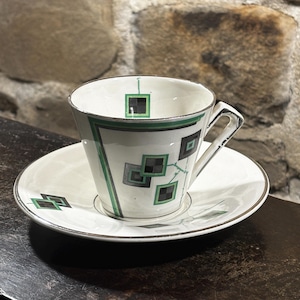 Könnte beinhalten: Weißes Tee-Set mit geometrischem Design in Schwarz und Grün. Die Tasse und die Untertasse haben einen silbernen Rand. Die Tasse hat einen Henkel und eine ausgestellte Form. Das Design umfasst Quadrate und Linien.