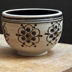 May include: A ceramic bowl with a beige exterior and a dark interior. The bowl features a repeating floral pattern in dark brown. The rim and base are also dark brown. The bowl is sitting on a wooden surface.