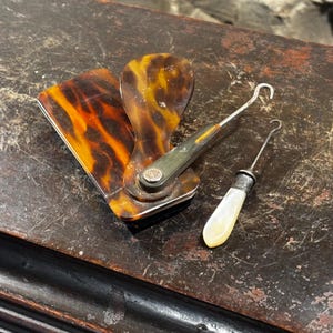 May include: A set of two tortoiseshell and silver button hook tools. One tool has a long handle and a hook on the end. The other tool has a shorter handle and a hook on the end. The tools are on a dark brown wooden surface.