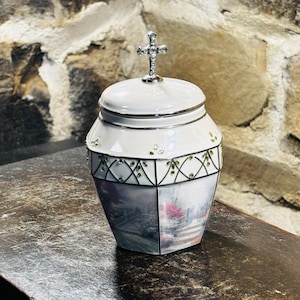 May include: A white ceramic urn with a silver rim and a cross-shaped lid handle. The urn features a painted landscape scene and is decorated with green accents. The urn is likely used for memorial purposes.