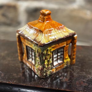 Puede incluir: Una caja de baratijas de cerámica con forma de cabaña con tejado de paja. La caja presenta un diseño detallado con ventanas y una chimenea. La paleta de colores incluye tonos de marrón, amarillo y verde, creando una estética rústica.