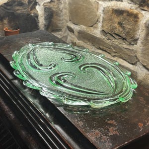 Könnte beinhalten: Ein hellgrünes ovales Serviertablett aus Glas mit einem dekorativen Wirbeldesign. Das Tablett hat einen gewellten Rand und besteht aus transparentem Glas. Das Tablett steht auf einer dunklen Holzoberfläche.