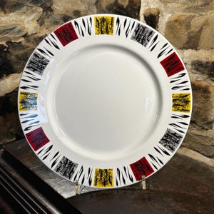 Może przedstawiać: Biały ceramiczny talerz z kolorowym geometrycznym wzorem. Obrzeże posiada naprzemienne kwadraty w kolorze czerwonym, żółtym i szarym, z czarnymi zakrzywionymi liniami. Talerz jest prezentowany na przezroczystym stojaku na tle kamienia.