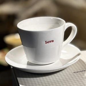 Puede incluir: Taza de café y platillo de cerámica blanca con la palabra "Love" impresa en rojo en el lateral de la taza.