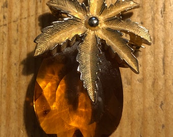 Op de afbeelding: Een broche van goudkleurige metaal met een grote amberkleurige glazen steen en een palmboomontwerp.