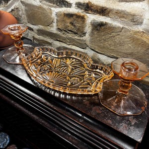 Könnte beinhalten: Ein Set aus vintage pfirsichfarbenen Glaskerzenhaltern und einer dekorativen Schale. Die Schale hat einen gewellten Rand und ein detailliertes Blumen- und geometrisches Muster. Die Kerzenhalter haben einen Sockel und eine sechseckige Oberseite.