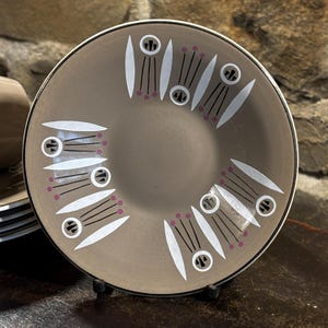 Könnte beinhalten: Ein brauner Keramikteller mit weißem und schwarzem Rand. Der Teller hat ein sich wiederholendes Muster aus weißen, tropfenförmigen Designs mit schwarzen Linien und kleinen rosa Punkten. Der Teller wird auf einem schwarzen Metallständer präsentiert.