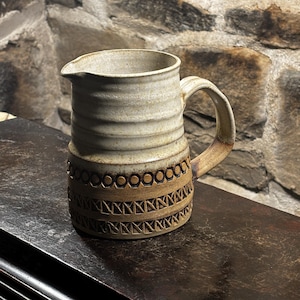 以下が含まれることがあります： 白地に茶色の幾何学模様が施された白い陶器のピッチャー。ピッチャーには持ち手と注ぎ口があります。