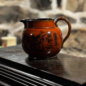 Puede incluir: Una jarra de cerámica marrón con asa negra y una escena en blanco y negro de personas y edificios. La jarra está sobre una superficie de madera oscura.