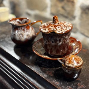 Puede incluir: Un juego de tres tazas de té de cerámica marrón y blanca con un patrón floral. Las tazas tienen una tapa y un platillo. Las tazas están sobre una superficie de madera oscura.