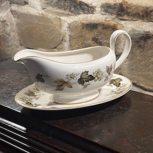 Può includere: Salsiera bianca con piattino abbinato. La salsiera ha un beccuccio curvo e un manico. Il set è decorato con un motivo floreale nei toni del verde e dell'oro. I bordi sono rifiniti in oro.