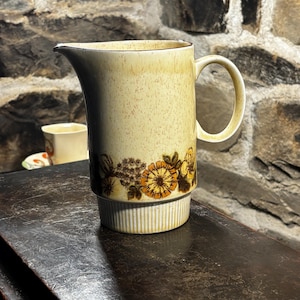 Könnte beinhalten: Vintage beige Keramikkrug mit floralem Design in Braun- und Orangetönen. Der Krug hat einen Henkel und einen Ausguss. Der Sockel hat ein geripptes Design. Er wird wahrscheinlich zum Servieren von Getränken verwendet.