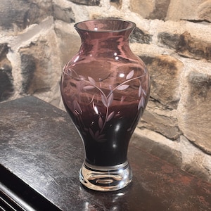 Puede incluir: Un jarrón de vidrio decorativo con un esquema de color degradado, que transiciona de una base púrpura oscura a un malva más claro en la parte superior. El jarrón presenta un diseño de hojas grabado. La base es de vidrio transparente.