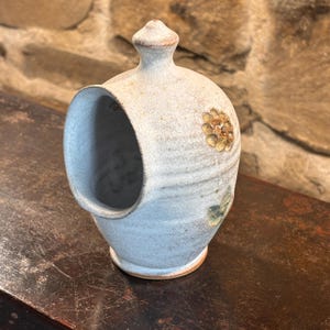 May include: A light blue ceramic salt pig with a wide, open side aperture and a small knobbed lid on top. The exterior features a textured, speckled finish with a small, embossed floral design in muted yellow and green tones on the front surface.