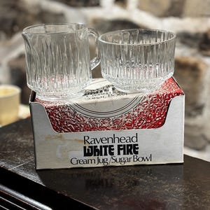 May include: A set of clear glass cream jug and sugar bowl with a textured design. The set is packaged in a white box with red and white text that reads "Ravenhead White Fire Cream Jug/Sugar Bowl".