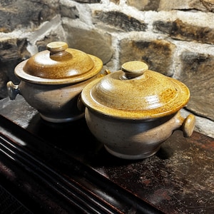 Puede incluir: Dos ollas de cerámica hechas a mano con tapas y asas. Las ollas son de color beige moteado con un borde y tapa marrón más oscuro. Las tapas tienen un pequeño pomo en la parte superior. Las ollas están sobre una superficie de madera oscura.