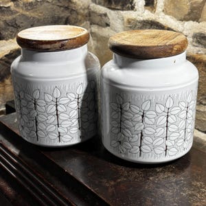 May include: Two white ceramic canisters with wooden lids. Each canister features a black and white leaf pattern. The canisters are cylindrical with a slightly rounded shape. The lids are round and made of light brown wood.