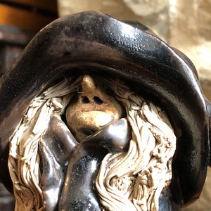 Peut inclure: Une figurine en céramique d'une femme aux longs cheveux blancs, portant un chapeau noir et une cape noire. Son visage est visible et elle a un grand nez.